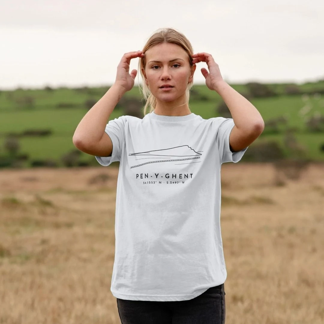Someone in a field wearing a white T-shirt with a line drawing of Pen-y-ghent